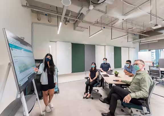 A group of employees wearing  masks are in the conference room with one of employee presenting